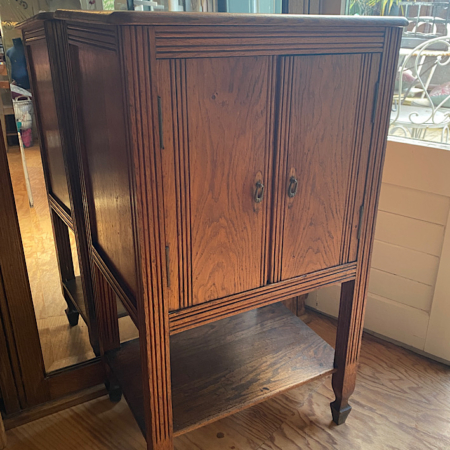 Un Meuble d’Appoint 1940 VENDU à deux portes et une étagère inférieure repose sur un plancher en bois près d’une fenêtre, dégageant un charme rétro rappelant la brocante 93. Brocante 93