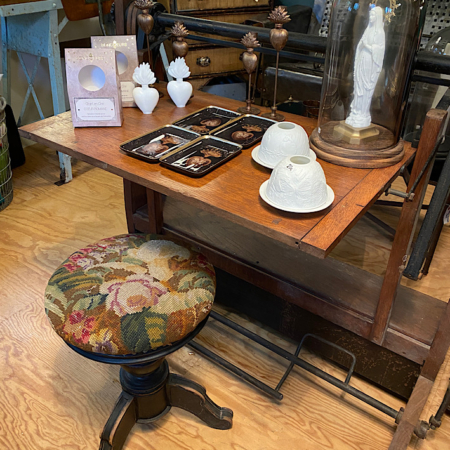 Vitrine vintage avec divers objets de décoration, tabouret à motifs floraux et Bureau cadastral XXème sur un plancher de bois. Cette charmante scène brocante 93 capture parfaitement l'attrait intemporel du style rétro. Brocante 93