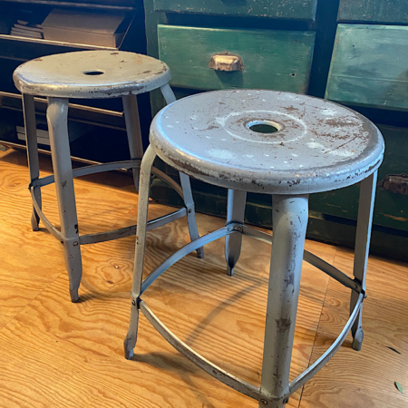 Deux Tabourets Nicolle 1960 aux assises rondes et trouées au centre, posés sur un plancher de bois contre des armoires vertes vintage, dégagent une charmante ambiance rétro rappelant les trésors de brocante 93. Brocante 93