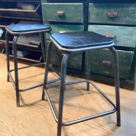 Deux tabourets de bar vintage Tabourets Industriels 1960 avec assises et dossiers noirs reposent sur un plancher en bois, devant des tiroirs verts. Brocante 93