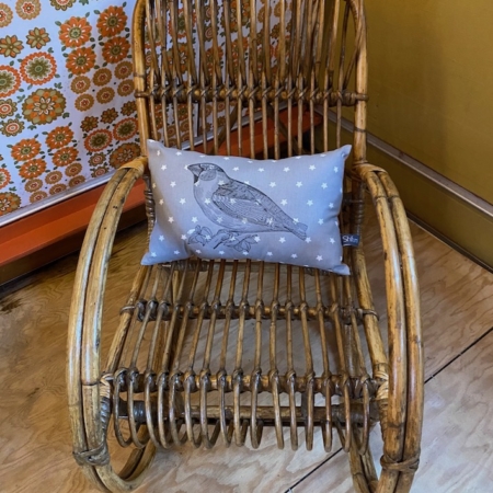 La Chaise à Bascule Enfant 1960 VENDU, ornée d'un coussin douillet mettant en valeur une illustration d'oiseau, adossée à un mur à motifs floraux, ajoute une charmante touche rétro à votre décoration intérieure. Brocante 93