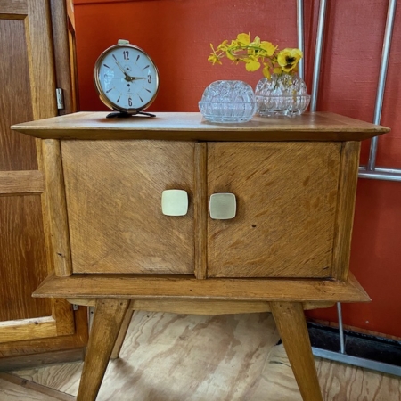 Une Table de Chevet rétro 1960 VENDU, comportant des portes, ornée d'une horloge classique et de fleurs jaunes dans des vases en verre sur le dessus, posée contre un mur rouge. Brocante 93