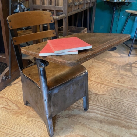 Un Bureau Américain Enfant 1950 VENDU, au charme vintage, présente un bureau d'école en bois avec une chaise attachée et un livre rouge posé sur la surface du bureau, capturant l'essence de la brocante classique 93. Brocante 93