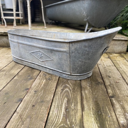 Une baignoire vintage en métal galvanisé "Baignoire Enfant 1950 VENDU" repose sur une terrasse en bois, accompagnée d'une autre grande baignoire en métal et de divers meubles d'extérieur en arrière-plan. Brocante 93