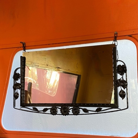 Miroir Fer Forgé 1950 avec un cadre floral orné suspendu sur un mur orange par deux chaînes. Brocante 93