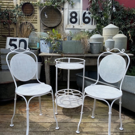 Deux chaises en métal blanc et une petite table ronde du Salon de Jardin 1950 VENDU implantées dans un jardin rustique au décor et aux plantes vintage évoquent une charmante ambiance rétro. Brocante 93