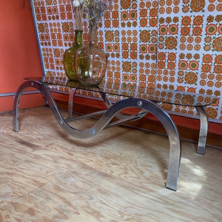 Banc métallique moderne vintage, connu sous le nom de Table Basse 1970 VENDU, doté d'un plateau en verre sur lequel sont posés des vases de fleurs, posé contre un mur à motifs floraux et un plancher en bois. Brocante 93