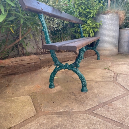 Le Banc de Jardin 1940 VENDU, avec son assise en bois patiné et ses pieds en fer vert, repose sur un patio en pierre, entouré de plantes feuillues et de hautes jardinières, donnant à l'espace une charmante ambiance rétro. Brocante 93