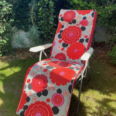 La chaise longue d'extérieur Transat de Jardin 1970, dotée d'un coussin floral rouge et rose vibrant, est posée sur une pelouse herbeuse avec de la verdure en arrière-plan, dégageant une charmante ambiance vintage. Brocante 93