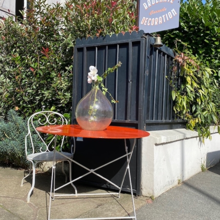 Une Table de Jardin 1970 rouge vif VENDU avec un vase transparent et des fleurs blanches, accompagnée d'une chaise en métal blanc et d'une pancarte "Brocante 93" sur un trottoir ensoleillé, ajoutant une touche de charme vintage. Brocante 93