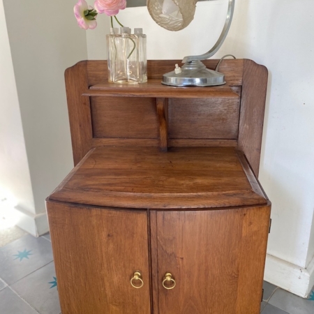 Une **Table de Chevet 1940 VENDU** aux poignées en laiton, ornée d'une lampe vintage et de fleurs roses dans des vases sur le dessus. Le sol présente des carreaux étoilés bleus, ajoutant au charme rétro du décor. Brocante 93