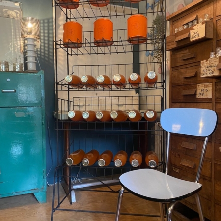 Présentoir Epicerie 1960 avec contenants orange et bouteilles marron, à côté d'un classeur bleu et d'une chaise bleu clair. Parfait pour ajouter une touche rétro à n'importe quel espace. Découvrez des pièces similaires chez Brocante 93. Brocante 93