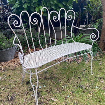 La Banquette Fer Forgé 1950 VENDU, au design de dossier orné et au charme rétro, est élégamment posée sur l'herbe dans un jardin entouré de plantes vertes et d'arbustes. Brocante 93