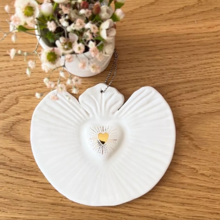 La décoration Coeur Sacré, une pièce en forme d'ange avec un coeur doré en son centre, repose sur une surface en bois patiné à côté d'un vase de petites fleurs, dégageant un charme vintage. Son esthétique brocante 93 renforce son élégance intemporelle. Brocante 93