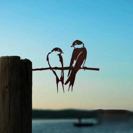 La Paire d'Hirondelles capture la silhouette de deux oiseaux perchés sur un poteau en bois avec un lac flou et un bateau en arrière-plan au coucher du soleil, évoquant un charme vintage rappelant les trésors de brocante rétro. Brocante 93