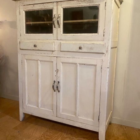 Le Buffet de Cuisine 1930 VENDU, un meuble en bois blanc usé avec des portes vitrées vintage en haut et des portes à panneaux fermés en bas, est placé contre un mur uni. Brocante 93