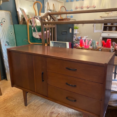 L'Enfilade Style Scandinave 1960, un buffet vintage en bois doté de trois tiroirs et d'une porte d'armoire, trône avec charme dans la brocante 93, entouré d'un ensemble d'objets et de meubles rétro. Brocante 93