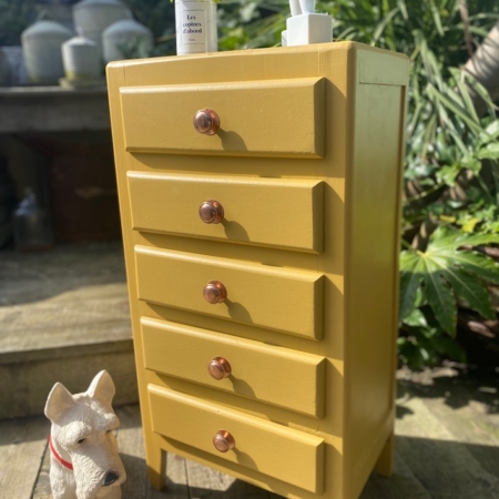 Le Semainier 1960 VENDU, une commode vintage jaune à cinq tiroirs, présente une petite plante et des produits de toilette sur le dessus. A côté, sur le plateau en bois, se trouve une charmante figurine de chien blanc, ajoutant une touche rétro. Brocante 93