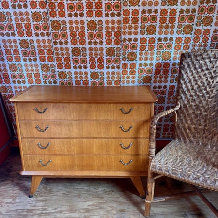 Commode Vintage 1960 à quatre tiroirs à côté d'une chaise en osier, sur fond de papier peint à motifs rétro. Brocante 93