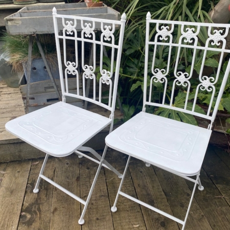 Deux chaises en métal blanc "Paire de Chaises 1960 VENDU" aux dossiers ornés reposent sur une terrasse en bois, entourée de plantes vertes luxuriantes, dégageant une charmante ambiance vintage. Brocante 93