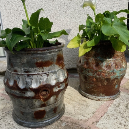 Deux jardinières en métal vintage "Paire de Fûts Industriels 1960" avec des plantes à feuilles vertes sont posées sur un patio en pierre près d'un mur blanc. Brocante 93