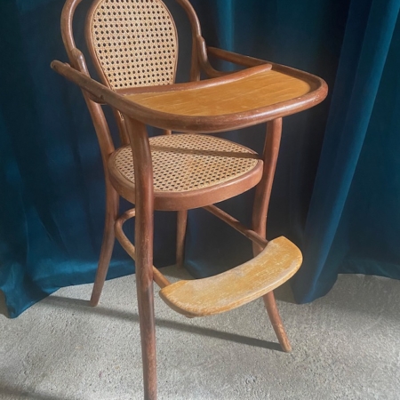 La Chaise haute bébé 1940 est une chaise haute vintage en bois avec dossier et assise en rotin, dotée d'un petit plateau et d'un repose-pieds, posée sur un rideau sombre. Cette pièce élégante dégage un charme rétro qui vous transporte dans une époque révolue de trésors de brocante 93. Brocante 93