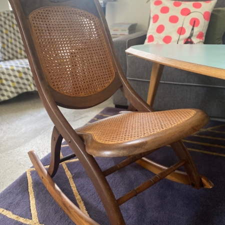 Une chaise à bascule Enfant 1940 avec une structure en bois vintage et une assise et un dossier tressés est posée sur un sol recouvert de moquette à côté d'une table basse et d'un canapé, évoquant une charmante ambiance rétro. Brocante 93