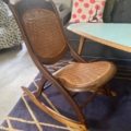 Une chaise à bascule Kids 1940 avec une assise et un dossier en rotin tressé repose sur un tapis à motifs près d'une table bleu sarcelle, parfaite pour le plaisir des adultes et des enfants.