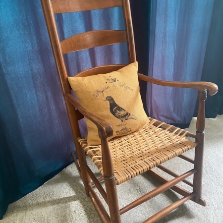 Le Rocking-Chair Américain 1930, une chaise à bascule vintage en bois avec une assise tressée et un coussin représentant une illustration de pigeon, posée sur un rideau bleu. Brocante 93