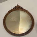 Un miroir ovale antique et rond du XIXème avec un cadre doré orné est accroché à un mur, montrant une ligne distincte où la moitié du verre semble sale et l'autre moitié est propre.