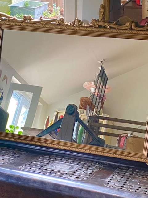 Miroir orné d'un cadre doré, le Miroir Ancien 1930, reflétant un intérieur chaleureux avec une porte ouverte et divers objets en arrière-plan.