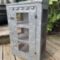 Une armoire en métal patiné avec des portes grillagées et des loquets repose sur une terrasse en bois, se fondant parfaitement dans l'environnement rustique, avec des plantes vertes en arrière-plan. La conception AUTO-DRAFT complète sans effort le cadre naturel.