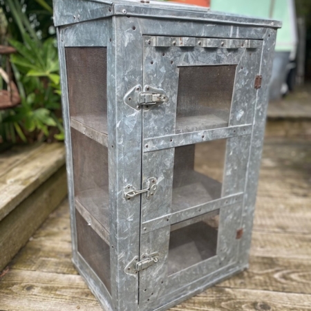 Une armoire extérieure en métal et en treillis, étiquetée AUTO-DRAFT, se dresse sur une terrasse en bois, avec des plantes et une clôture en bois en arrière-plan.