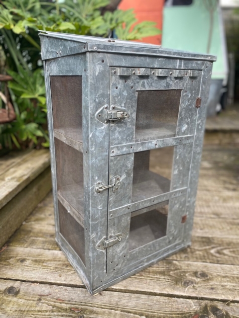 Une armoire extérieure en métal et en treillis, étiquetée AUTO-DRAFT, se dresse sur une terrasse en bois, avec des plantes et une clôture en bois en arrière-plan.