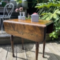 Table à rabats des années 1940 à décor floral et vases remplis de fleurs, posée sur une terrasse en bois entourée de verdure. Brocante 93