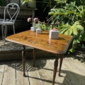 La Table à rabats 1940, ornée de petites plantes en pot et de fleurs, trône gracieusement sur une terrasse en bois dans un décor de jardin extérieur. Brocante 93