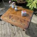 La Table à rabats 1940, dotée d'un élégant design en bois aux motifs floraux, est complétée par un décor carré blanc et un petit vase rempli de fleurs roses, et repose gracieusement sur une terrasse en bois. Brocante 93