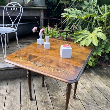 Une Table à rabats 1940 aux motifs floraux supporte trois vases blancs remplis de fleurs. Une chaise blanche et de la verdure en arrière-plan contribuent à l'élégance intemporelle de la scène. Brocante 93