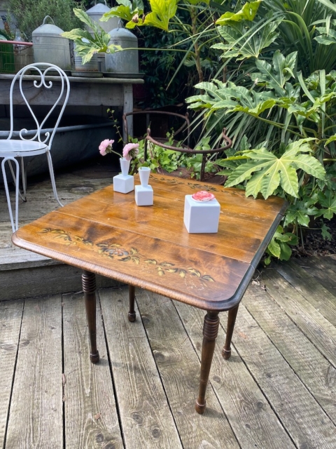 Une Table à rabats 1940 aux motifs floraux supporte trois vases blancs remplis de fleurs. Une chaise blanche et de la verdure en arrière-plan contribuent à l'élégance intemporelle de la scène. Brocante 93