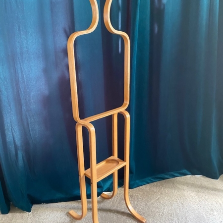 Valet de Chambre 1970, un porte-manteau abstrait en bois de forme humaine avec une étagère intégrée, se dresse élégamment contre un rideau bleu sarcelle foncé. Brocante 93