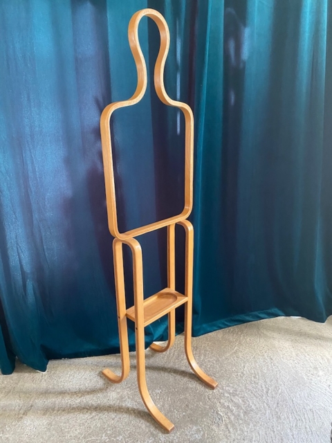 Valet de Chambre 1970, un porte-manteau abstrait en bois de forme humaine avec une étagère intégrée, se dresse élégamment contre un rideau bleu sarcelle foncé. Brocante 93