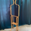 Une sculpture abstraite en bois ressemblant à une forme humaine simplifiée se dresse devant un rideau bleu sarcelle foncé, évoquant l'élégance minimaliste du Valet de Chambre 1970. Brocante 93