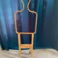 Le Valet de Chambre 1970, un meuble en bois évoquant une silhouette humaine simplifiée, se dresse sur un fond de rideau bleu sarcelle foncé. Son design intemporel évoque l'élégance des années 1970. Brocante 93