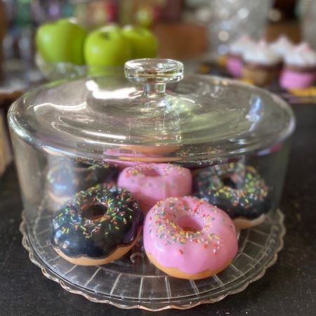 Beignets avec glaçage rose et noir et pépites colorées sous un dôme en verre sur un comptoir, avec des pommes, des cupcakes et une machine à pression automatique à proximité. Brocante 93