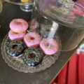 Assiette avec des beignets glacés roses et noirs avec des pépites sous un dôme en verre, à côté d'un AUTO-DRAFT de cupcakes colorés en arrière-plan. Brocante 93