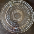 Une assiette ronde en verre transparent avec un motif en étoile complexe et un bord festonné, rappelant les années 1950, posée sur une surface sombre. Parfaite comme pièce d'appoint Cloche à Gâteaux 1950. Brocante 93