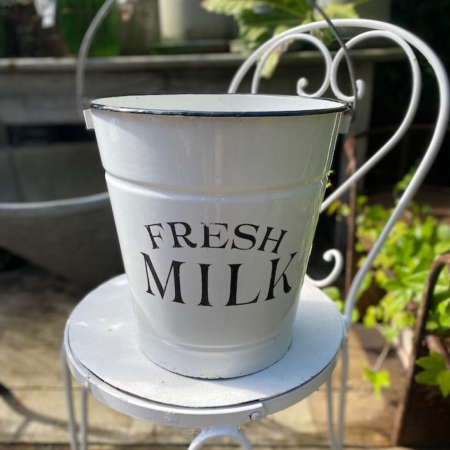 Un charmant Seau à Lait USA 1950 en métal blanc, étiqueté « Fresh Milk », trône gracieusement sur une chaise décorative en métal blanc dans un décor de jardin serein, rappelant une campagne américaine des années 1950. Une touche d'élégance rustique, ce seau à lait ajoute un charme nostalgique à la scène pittoresque. Brocante 93