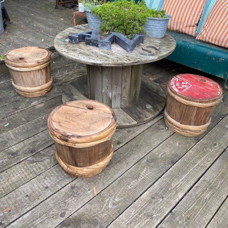 Espace extérieur avec une table ronde en bois et trois tabourets tonneaux sur une terrasse en bois, dégageant une esthétique japonaise. Des plantes en pot et la bouteille de Fût à Saké Japonais 1960 sur la table renforcent l'ambiance sereine. Brocante 93