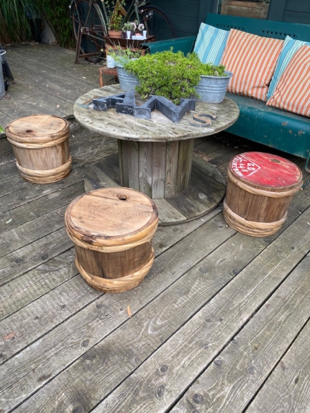 Espace extérieur avec une table ronde en bois et trois tabourets tonneaux sur une terrasse en bois, dégageant une esthétique japonaise. Des plantes en pot et la bouteille de Fût à Saké Japonais 1960 sur la table renforcent l'ambiance sereine. Brocante 93