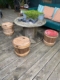 Espace extérieur avec une table ronde en bois et trois tabourets tonneaux sur une terrasse en bois, dégageant une esthétique japonaise. Des plantes en pot et la bouteille de Fût à Saké Japonais 1960 sur la table renforcent l'ambiance sereine. Brocante 93